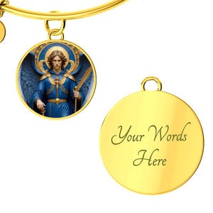 May include: Gold-toned bangle bracelet with two round charms. One charm features a detailed illustration of an angel in blue robes with blue wings. The other charm is blank with the words "Your Words Here" in script.