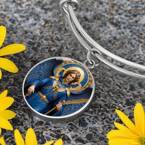 May include: A silver-toned bangle bracelet featuring a round pendant with a detailed illustration of an angel. The angel is depicted with blue wings and a blue robe, holding a golden staff. The background is a mix of blue and gold.