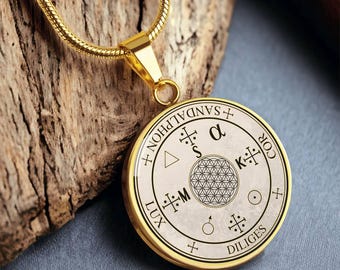 May include: Gold-toned pendant necklace with a circular charm. The charm features a cream-colored background with black symbols and text, including a geometric pattern. The necklace is displayed on a wooden surface.