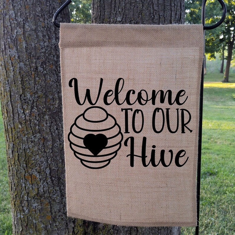 May include: A rectangular burlap garden flag with the words "Welcome TO OUR Hive" in black script. A black beehive graphic with a heart is centered below the word "Welcome". The flag is hanging on a black metal stand.