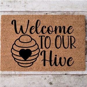 May include: A rectangular doormat made of natural coir fiber. The mat features the words "Welcome TO OUR Hive" in a black, stylized font. A black heart is inside a beehive graphic.