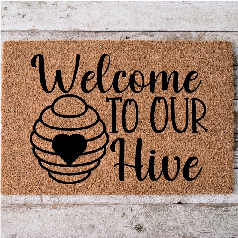 May include: A rectangular doormat made of natural coir fiber. The mat features the words "Welcome TO OUR Hive" in a black, stylized font. A black heart is inside a beehive graphic.