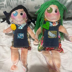 May include: Two handmade fabric dolls with colourful clothing and yarn hair. One doll has black hair and blue eyes, the other has green hair and brown eyes.