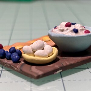 May include: Miniature food arrangement featuring a brown wooden serving board. It holds a light blue bowl of cream with red and blue berries, a yellow dish of white objects, and clusters of red and blue berries.