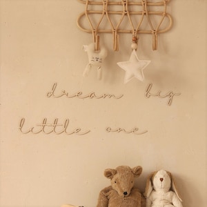 Può includere: Un set di decorazioni murali per la cameretta dei bambini con le parole "dream big little one" in corsivo. Il set include una stella di legno, un coniglietto e un appendiabiti da parete in rattan con ganci.