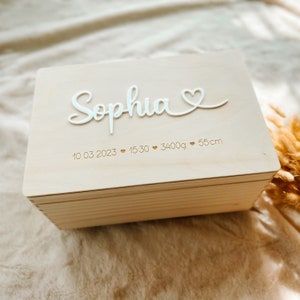Peut inclure: Un coffret souvenir en bois clair avec le nom "Sophia" en écriture blanche. La boîte comprend la date, l'heure, le poids (3400g) et la taille (55cm) d'un bébé. La boîte est fermée et posée sur une surface douce et de ton neutre.