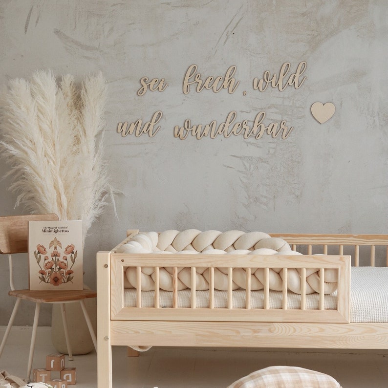 Op de afbeelding: Een houten bed met een witte gevlochten bumper en een muur met de woorden "sei frech wild und wunderbar" en een klein hart.