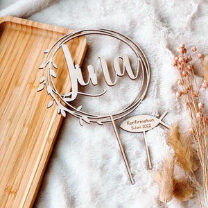 May include: Wooden cake topper with the name "Lina" in a cursive font, surrounded by a wreath of leaves. The topper also includes a small wooden fish with the text "Confirmation 5. Juni 2022".