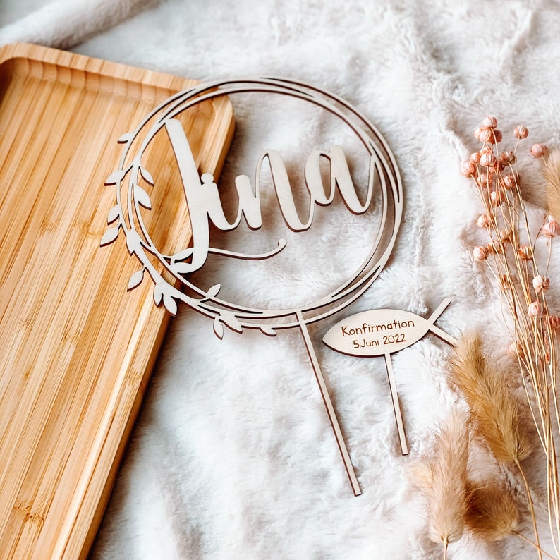 May include: Wooden cake topper with the name "Lina" in a cursive font, surrounded by a wreath of leaves. The topper also includes a small wooden fish with the text "Confirmation 5. Juni 2022".