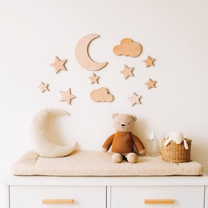 May include: A nursery scene featuring a beige changing pad on a white dresser. A plush teddy bear and a crescent moon pillow sit on the pad. Wooden stars, clouds, and a moon decorate the wall, creating a whimsical atmosphere.