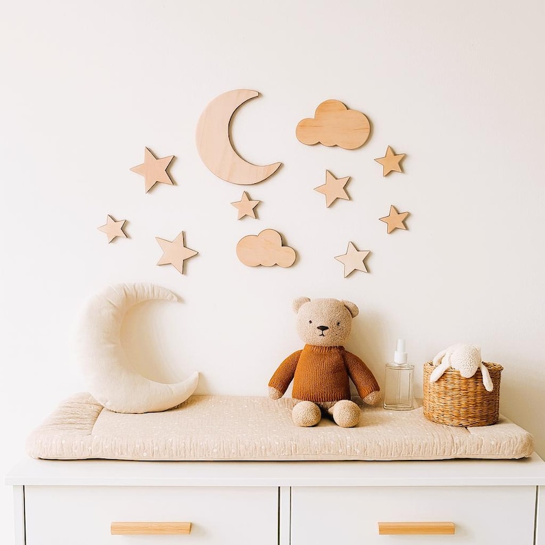 May include: A nursery scene featuring a beige changing pad on a white dresser. A plush teddy bear and a crescent moon pillow sit on the pad. Wooden stars, clouds, and a moon decorate the wall, creating a whimsical atmosphere.
