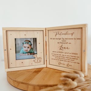 May include: A wooden box with a hinged lid, engraved with a message about a godparent. The box has a small photo frame with a picture of a baby girl. The text in the box reads: "Patenbrief Mit dem heutigen Tag &uuml;bernimmst du liebe Carolin die Patenschaft von Lena geboren am 18.10.2024 | getauft am 15.04.2025 Ich freue mich sehr, dass du meine Patentante bist, dass du mich auf meinem Lebensweg begleitest, mir mit Rat und Tat zur Seite stehst und mich an den H&auml;nden h&auml;ltst, damit ich nicht falle Deine Lena"