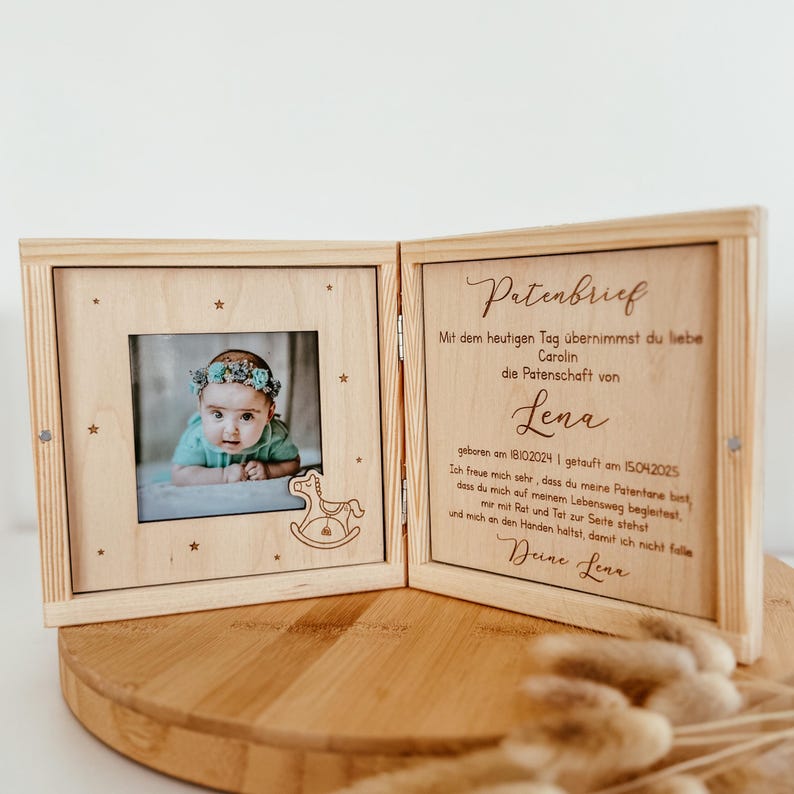 May include: A wooden box with a hinged lid, engraved with a message about a godparent. The box has a small photo frame with a picture of a baby girl. The text in the box reads: "Patenbrief Mit dem heutigen Tag &uuml;bernimmst du liebe Carolin die Patenschaft von Lena geboren am 18.10.2024 | getauft am 15.04.2025 Ich freue mich sehr, dass du meine Patentante bist, dass du mich auf meinem Lebensweg begleitest, mir mit Rat und Tat zur Seite stehst und mich an den H&auml;nden h&auml;ltst, damit ich nicht falle Deine Lena"