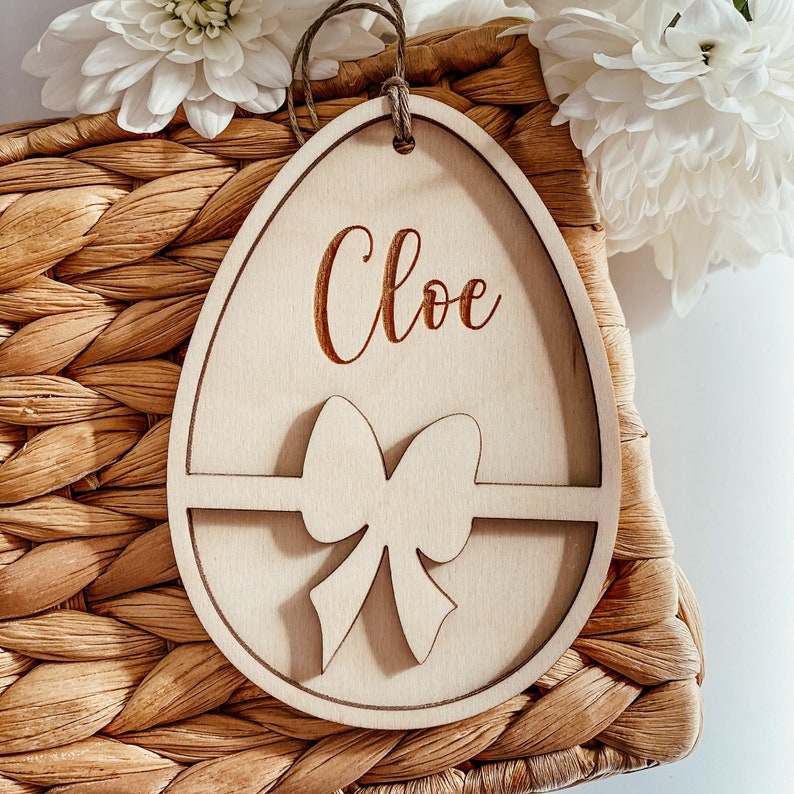 May include: A wooden egg-shaped ornament with a bow and the name "Chloe" engraved on it. The ornament is sitting on a woven basket.