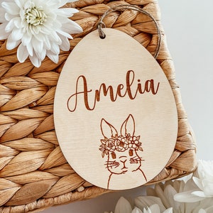 May include: A wooden Easter egg ornament with a bunny wearing a flower crown and the name Amelia engraved on it.
