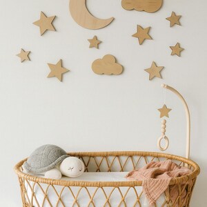 May include: A nursery scene featuring a wicker bassinet with a white sheet, a gray turtle plush, and a white plush toy. Wooden stars, clouds, and a crescent moon decorate the wall. A wooden mobile hangs above the bassinet.