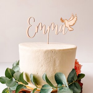 May include: A white cake with a wooden cake topper that says "Emma" with a dove holding an olive branch. The cake is decorated with greenery.