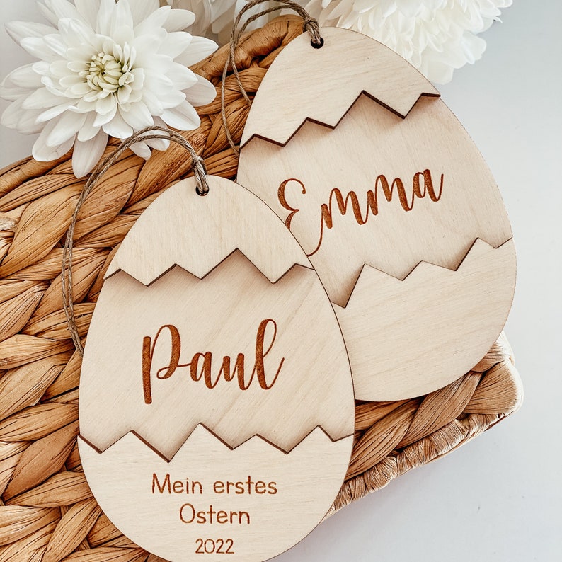 May include: Two wooden Easter egg ornaments with the names "Emma" and "Paul" engraved on them. The ornament with "Paul" also has the text "Mein erstes Ostern 2022" engraved on it.