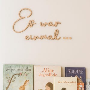 Könnte beinhalten: Wanddekoration aus Holz mit der Aufschrift "Es war einmal..." in Schreibschrift. Darunter eine Sammlung von Büchern mit Titeln wie "Wilgen Weisheiten" und "Alice Jugendliebe". Die Dekoration und die Bücher sind vor einer neutralen Wand angeordnet.