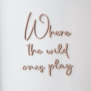 May include: Wooden wall art spelling out "Where the wild ones play" in a cursive font. The letters are a light brown colour and are mounted on a white wall. A decorative piece for a nursery or child's room.