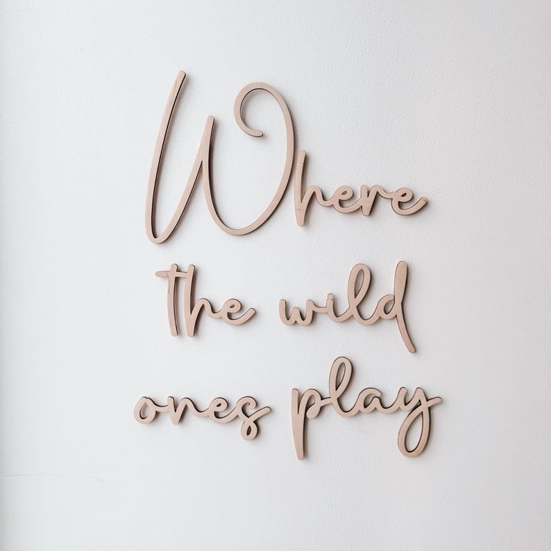 May include: Wooden wall art spelling out "Where the wild ones play" in a cursive font. The letters are a light brown colour and are mounted on a white wall. A decorative piece for a nursery or child's room.