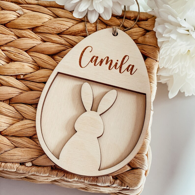 May include: A wooden Easter egg-shaped ornament with the name "Camila" engraved on the top and a cutout of a bunny rabbit on the bottom.