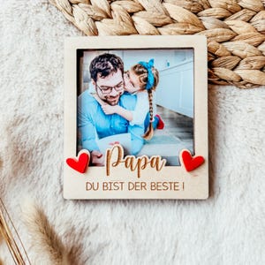 May include: A wooden photo frame with a picture of a father and daughter. The frame has two red hearts on the bottom and the text "Papa Du Bist Der Beste!"