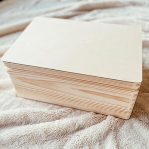 May include: A light-colored, rectangular wooden box with a lid. The box is made of natural wood and has visible grain patterns. The box is sitting on a soft, beige surface.