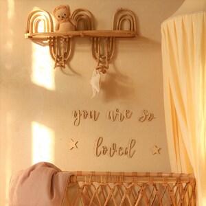 May include: A nursery room with a wicker crib, a wooden shelf with a stuffed animal, and a sign that reads "you are so loved" with two stars on either side.