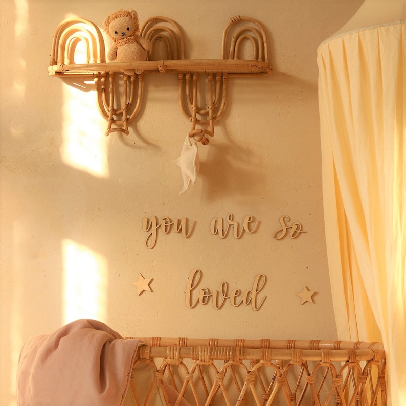 May include: A nursery room with a wicker crib, a wooden shelf with a stuffed animal, and a sign that reads "you are so loved" with two stars on either side.