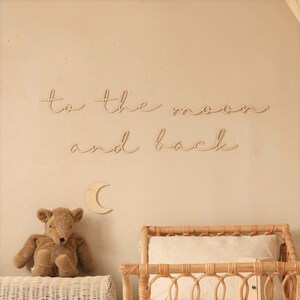 May include: Wooden wall decor featuring the phrase "to the moon and back" in cursive script. A small wooden crescent moon is positioned below the text. A teddy bear and a wicker crib are in the background.
