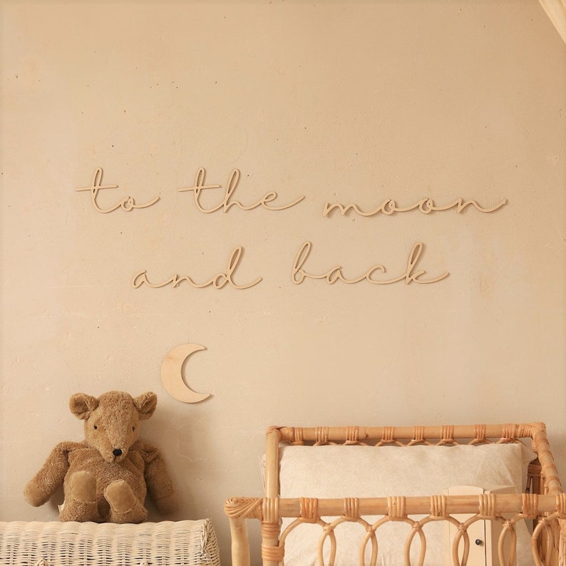 May include: Wooden wall decor featuring the phrase "to the moon and back" in cursive script. A small wooden crescent moon is positioned below the text. A teddy bear and a wicker crib are in the background.
