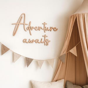 Op de afbeelding: Houten wanddecoratie met de tekst "Adventure awaits" in een sierlijk lettertype. Daaronder hangt een slinger van kleine, driehoekige vlaggetjes. Een beige luifel en kussens op de achtergrond creëren een bohemien sfeer.