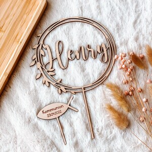 May include: Wooden cake topper with a circle wreath design and the name "Henry" in cursive. The topper also includes a small wooden fish with the text "Kommunion 23.04.2022".
