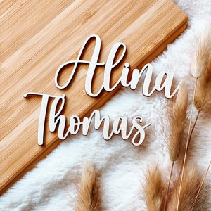 Puede incluir: Dos letreros de madera con los nombres "Alina" y "Thomas" est&aacute;n colocados sobre una superficie de madera marr&oacute;n claro. Los letreros est&aacute;n en una fuente cursiva y son de color blanco.