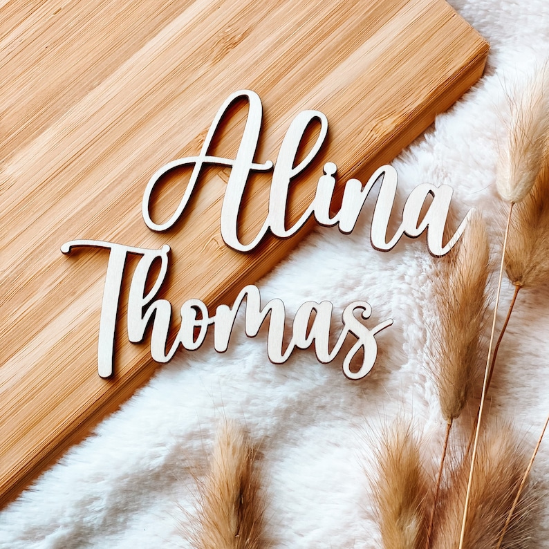 Puede incluir: Dos letreros de madera con los nombres "Alina" y "Thomas" est&aacute;n colocados sobre una superficie de madera marr&oacute;n claro. Los letreros est&aacute;n en una fuente cursiva y son de color blanco.