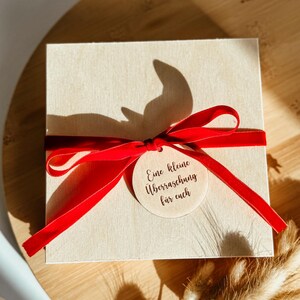 May include: A square, light-coloured wooden box tied with a vibrant red ribbon. A small, round wooden tag with the text "Eine kleine &Uuml;berraschung f&uuml;r euch" is attached to the ribbon. The box is sitting on a wooden surface.