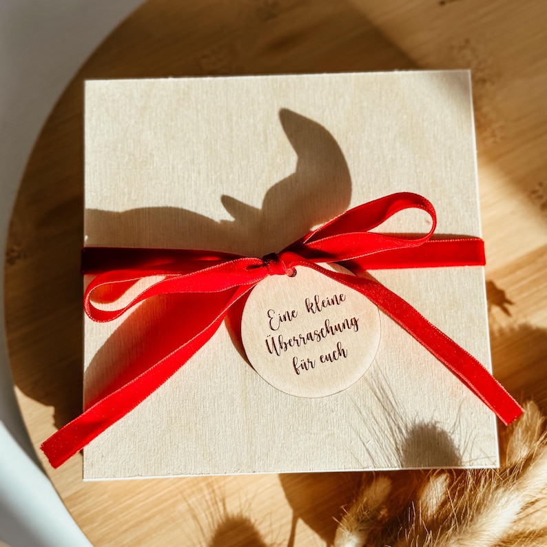 May include: A square, light-coloured wooden box tied with a vibrant red ribbon. A small, round wooden tag with the text "Eine kleine &Uuml;berraschung f&uuml;r euch" is attached to the ribbon. The box is sitting on a wooden surface.