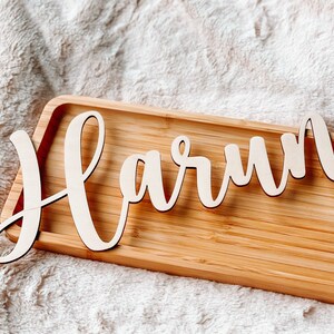 Puede incluir: Una bandeja de madera con el nombre "Harun" escrito en cursiva. La bandeja es de madera marr&oacute;n claro y tiene un acabado natural.