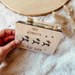 May include: A wooden Christmas gift card holder with a reindeer design and the German text "Frohe Weihnachten" and "F&uuml;r Julia". A ten-dollar bill is visible inside the holder.