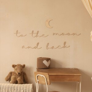 May include: Wooden wall decor with the phrase "to the moon and back" in cursive script. A small wooden crescent moon is above the text. A brown wicker basket with a white heart is on a wooden desk with a white chair.
