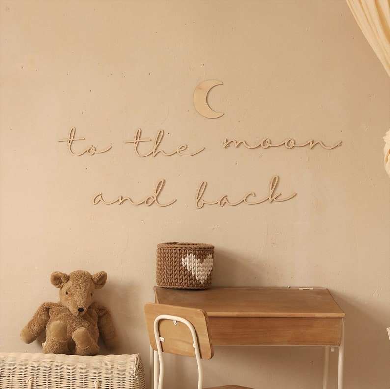 May include: Wooden wall decor with the phrase "to the moon and back" in cursive script. A small wooden crescent moon is above the text. A brown wicker basket with a white heart is on a wooden desk with a white chair.