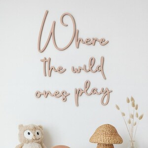May include: Soft pink wooden wall art spelling out "Where the wild ones play" in a cursive font. The wall art is mounted on a white wall. A stuffed owl, woven lamp, and dried flowers are in the foreground.