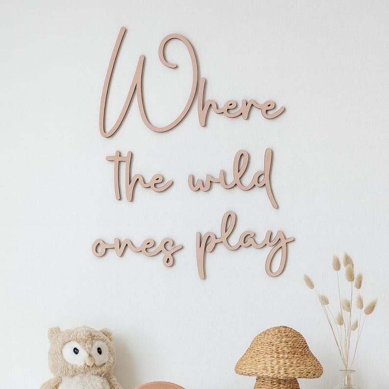 May include: Soft pink wooden wall art spelling out "Where the wild ones play" in a cursive font. The wall art is mounted on a white wall. A stuffed owl, woven lamp, and dried flowers are in the foreground.