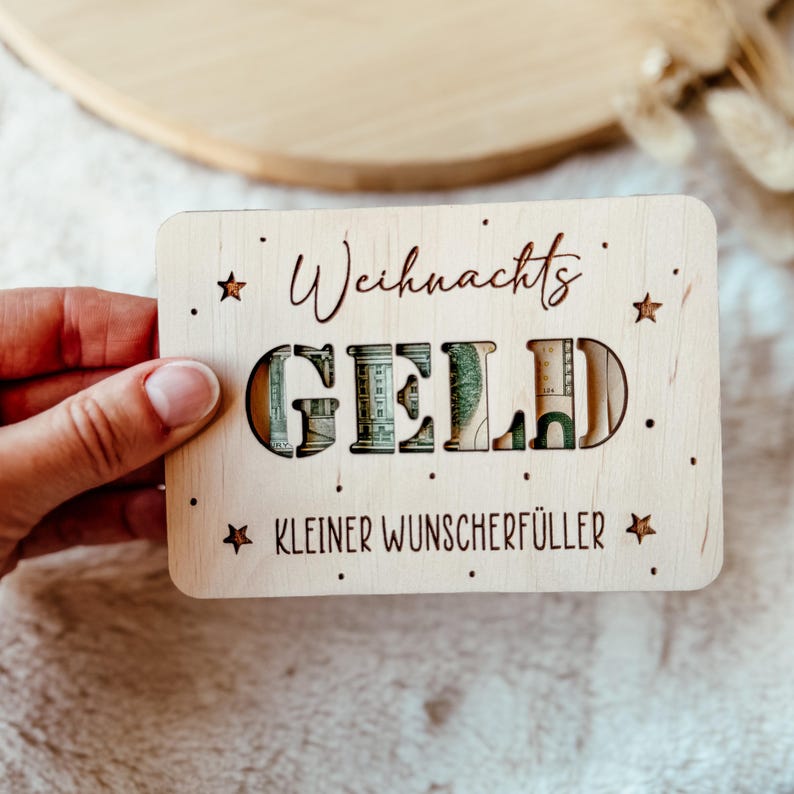 May include: A wooden gift card holder with the word "GELD" cut out, revealing money inside. The words "Weihnachts" and "Kleiner Wunscherf&uuml;ller" are written on the card, along with star and dot embellishments. The card is held by a hand.