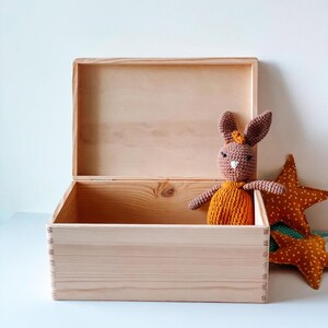 May include: An open, light-colored wooden box with a hinged lid. Inside sits a crocheted brown bunny wearing an orange dress. Two orange star-shaped pillows with white polka dots rest beside the box, suggesting a children's theme.