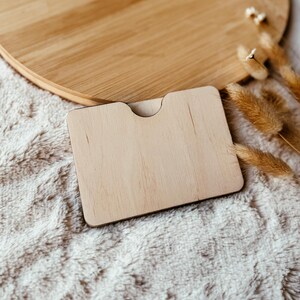 May include: A light-colored wooden card holder with a curved top cutout, resting on a soft, white, textured surface. A round wooden tray is in the background. Dried wheat stalks are to the right.