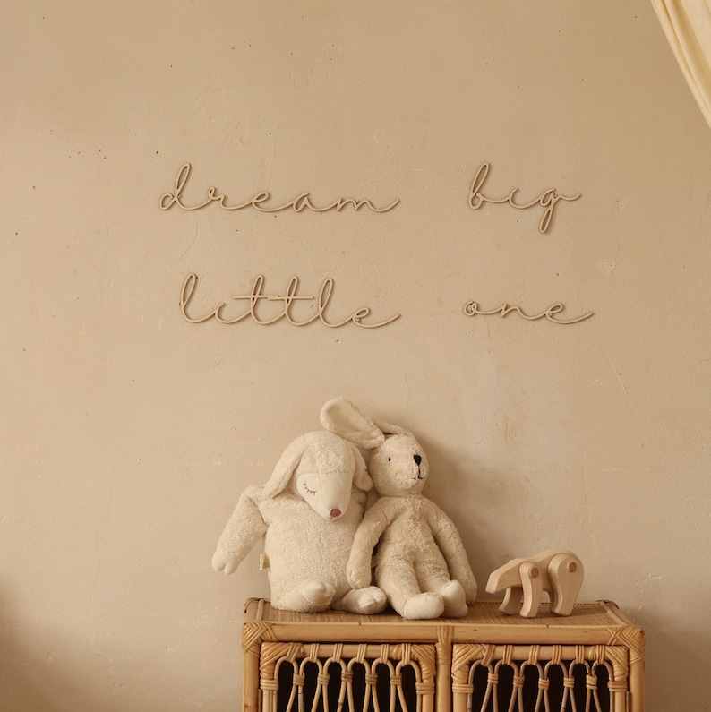 May include: Wooden wall decor with the words "dream big little one" in a cursive font. Two plush toys, a white bunny and a white lamb, are sitting on a wicker shelf.