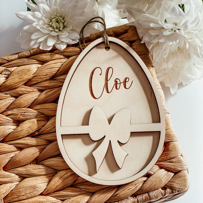 May include: A wooden Easter egg-shaped ornament with a bow and the name "Chloe" engraved on it. The ornament is sitting on a woven basket.