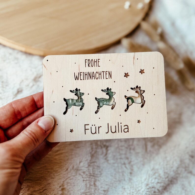 May include: A wooden Christmas card with the words "Frohe Weihnachten" and "F&uuml;r Julia" printed on it. The card features three reindeer cutouts with patterned designs. Small star and dot accents are scattered around the text and reindeer.